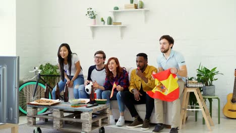 Grupo-multiétnico-de-amigos-deportes-aficionados-con-banderas-españolas-viendo-el-Campeonato-de-fútbol-en-la-televisión-juntos-en-casa-y-animar-para-arriba-equipo-favorito