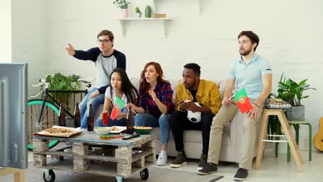 Grupo-multiétnico-de-amigos-deportes-aficionados-con-banderas-portuguesas-viendo-el-Campeonato-de-fútbol-en-la-televisión-juntos-en-casa-y-animar-para-arriba-equipo-favorito