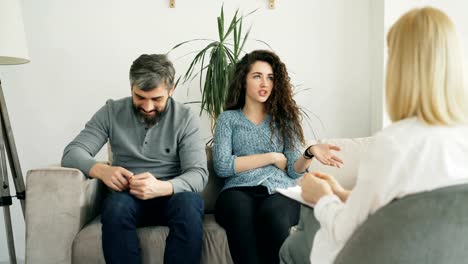 Young-couple-visiting-professional-psychologist-office.-Curly-woman-angrily-arguing-and-talking-about-her-boyfriend-to-marriage-counselor