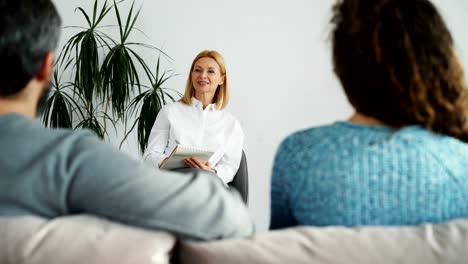 Female-professional-psychologist-talking,-listening-and-writing-notes-in-notebook-while-young-arguing-couple-visit-her-office-indoors