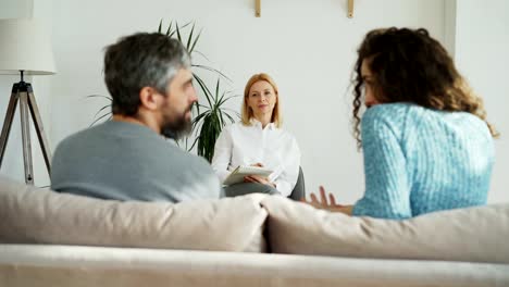 Female-experienced-psychoanalyst-listening-and-writing-notes-while-married-angry-couple-arguing-and-quarreling-during-visit-professional-psychologist-and-marriage-counselor-office