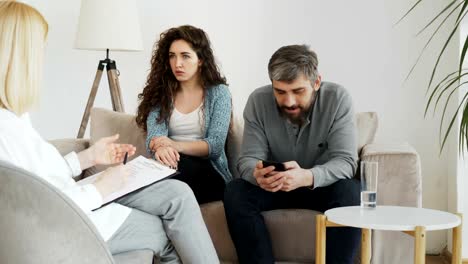 Female-professional-psychologist-listening-and-writing-notes-while-young-woman-arguing-and-her-boyfriend-surfing-in-smartphone-during-visit-psychotherapy-office-indoors