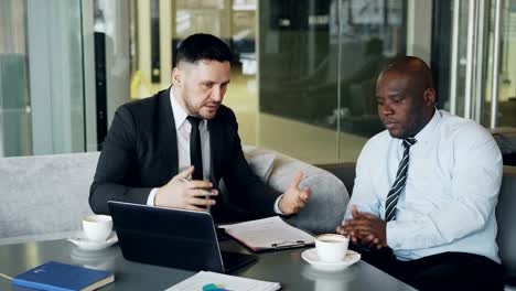 Angry-businessman-in-black-business-suit-criticizes-severely-his-african-american-employee-during-meeting-in-modern-cafe.-Irritated-boss-gesticulates-emotionally-and-agressively-shout