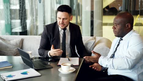 Gereizte-Bussinessman-im-Anzug-Schelte-verärgert-seine-afroamerikanische-Mitarbeiter-im-Hemd-im-modernen-Café.-Wütend-ceo-schlägt-die-Tabelle-mit-der-Hand-nervös