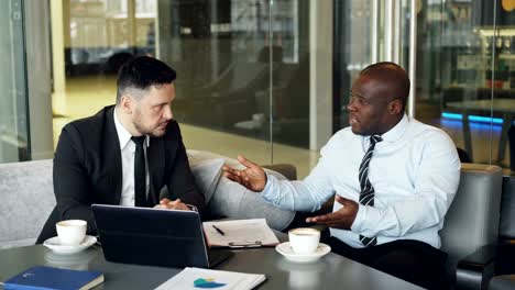 Empresario-enojado-en-la-camisa-una-corbata-critica-severamente-su-compañero-durante-la-reunión-en-el-café-moderno.-Jefe-afroamericano-irritada-emocionalmente-gesticula-y-habla-agresivamente