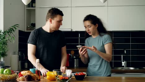 schöner-junger-kaukasischer-Mann-am-Tisch-Schneiden-von-Gemüse-stehen-und-versucht-Stück-gelbe-Paprika,-seine-Freundin-kommt-wütend-halten-Handys,-Mann-lachen