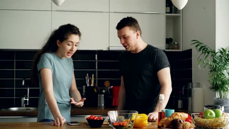 joven-mujer-rizada-mostrando-algo-desagradable-en-el-teléfono-del-esposo-mientras-él-se-cocina,-gritan-y-el-pelearse,-hombre-enojado-e-irritado