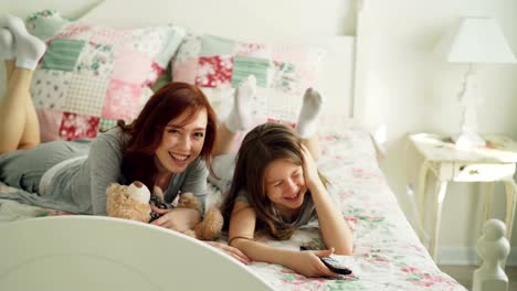 Beautiful-happy-mother-with-little-daughter-watching-cartoon-movie-on-TV-and-laughing-while-lying-on-bed-at-home-in-the-morning-in-cozy-bedroom