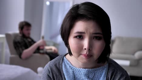 Portrait-of-a-sad-depressed-Asian-girl,-drunk-husband-in-the-background,-looking-at-the-camera.-50-fps