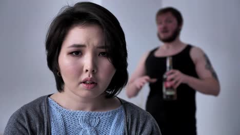 Portrait-of-a-sad-depressed-Asian-girl,-drunk-husband-in-the-background-swears,-alcohol,-conflict,-looks-at-the-camera.-50-fps