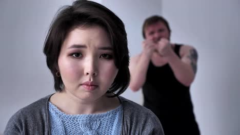 Portrait-of-a-sad-depressed-Asian-girl,-drunk-husband-in-the-background-is-grieving,-alcohol,-violence,-looks-at-the-camera.-60-fps