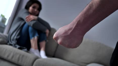 Concept-of-domestic-violence-in-family,-male-fist-afraid-Asian-woman-sitting-on-the-couch-in-the-background-50-fps