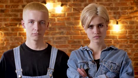 Young-serious-couple-of-hipsters-is-watching-at-camera,-girl-is-crossinf-her-arms,-brick-background