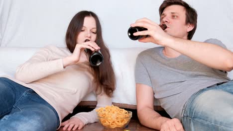 Pareja-joven-hombre-y-mujer-mienten-en-el-piso-en-casa-comiendo-patatas-fritas-y-bebiendo-cerveza-y-hablando.