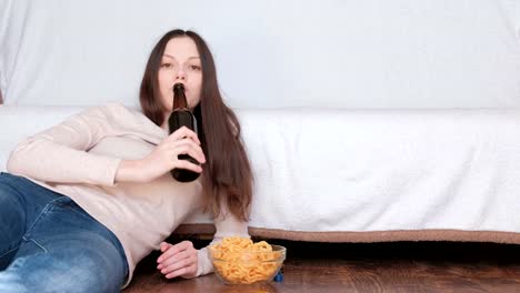 Mujer-hermosa-joven-bebiendo-una-cerveza-y-comiendo-chips-tendido-en-el-piso-cerca-de-la-cama.