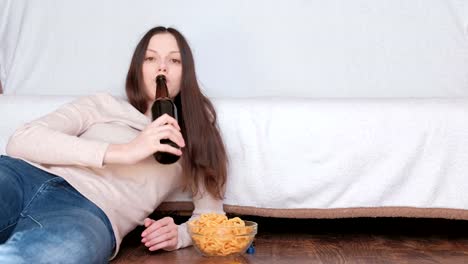 Young-beautiful-woman-drinking-a-beer-and-eating-chips-laying-on-the-floor-near-the-couch.