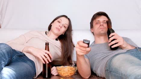 Pareja-de-jóvenes-hombre-y-mujer-comiendo-fritas-bebiendo-cerveza-y-viendo-la-TV,-cambio-de-canal.-Hablando-y-sonriendo