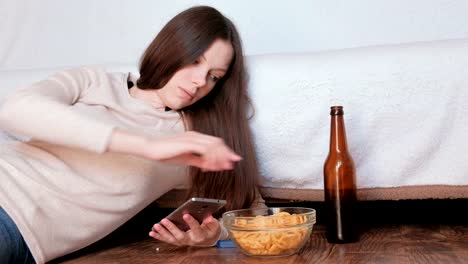 Sad-young-beautiful-brunette-woman-typing-a-message-on-the-mobile-phone,-drinks-beer-and-eats-chips.