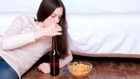 Morena-hermosa-joven-bebiendo-una-cerveza-y-comiendo-chips-tendido-en-el-piso-cerca-del-sofá-y-viendo-la-televisión.