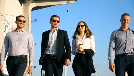 Group-of-young-happy-business-partners-walking-in-city-street-to-airport-terminal.-Colleagues-being-on-his-way-to-business-trip.-Portrait-of-smiling-business-people-stepping-and-talking.-Slow-motion-Close-up