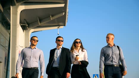 Group-of-young-happy-business-partners-walking-in-city-street-to-airport-terminal.-Colleagues-being-on-his-way-to-business-trip.-Portrait-of-smiling-business-people-stepping-and-talking.-Slow-motion-Close-up