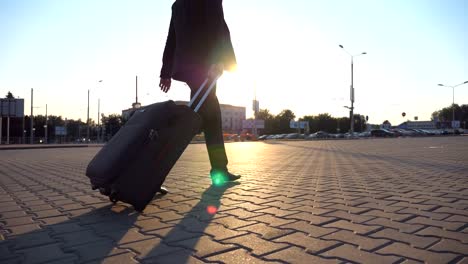 Profil-von-erfolgreicher-Geschäftsmann-im-schwarzen-Anzug-zu-Fuß-mit-seinem-Gepäck-auf-städtischen-Straße-bei-Sonnenuntergang.-Zuversichtlich-junger-Mann-geht-zum-Flughafenterminal-und-ziehen-Koffer-auf-Rädern.-Nahaufnahme-Seitenansicht