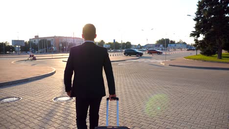 Rear-view-of-unrecognizable-young-businessman-walking-to-airport-terminal-and-pulling-suitcase-on-wheels-at-sunset.-Successful-male-business-person-going-with-his-luggage-on-city-street.-Close-up