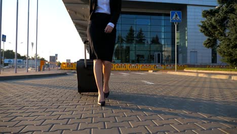 Young-woman-walking-near-airport-parking-with-her-luggage-at-sunset-time.-Female-legs-in-high-heels-stepping-at-sidewalk.-Business-woman-going-with-her-suitcase-on-urban-street.-Slow-motion-Close-up