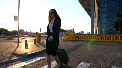 Junge-Frau,-die-in-der-Nähe-parken-am-Flughafen-mit-ihrem-Gepäck-bei-Sonnenuntergang.-Business-Frau-geht-mit-ihrem-Koffer-entlang-Stadtstraße.-Sonne-Streulicht-im-Hintergrund.-Reisekonzept.-Slow-Motion-hautnah