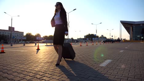 Mujer-en-calzado-tacón-alto-caminando-cerca-de-aparcamiento-en-el-aeropuerto-con-su-equipaje-en-el-atardecer.-Empresaria-con-su-maleta-a-lo-largo-de-la-calle-de-la-ciudad.-El-sol-de-la-llamarada-en-el-fondo.-Lenta-de-cerca