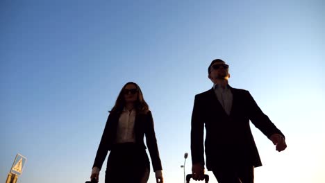 Business-man-and-woman-going-to-the-airport-with-their-luggage.-Young-businessman-carrying-suitcase-on-wheels-and-walking-with-his-female-colleague-to-terminal-hall.-Trip-or-travel-concept.-Low-angle-view-Slow-motion