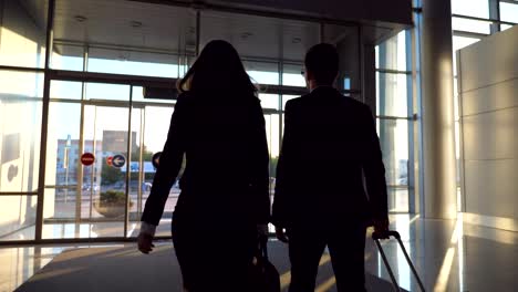 Business-man-and-woman-with-luggage-going-from-the-airport-to-city-street.-Follow-to-young-businessman-carrying-suitcase-on-wheels-and-walking-with-his-female-colleague-from-terminal-hall.-Rear-view-Close-up