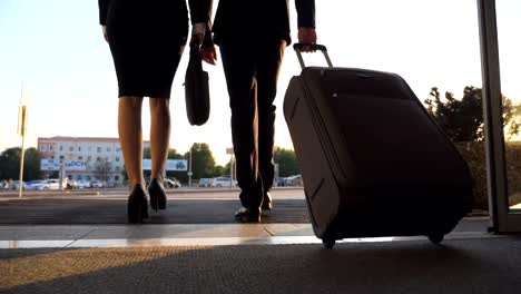 Business-man-and-woman-with-luggage-going-from-the-airport-to-city-street.-Follow-to-young-businessman-carrying-suitcase-on-wheels-and-walking-with-his-female-colleague-from-terminal-hall.-Rear-view-Close-up