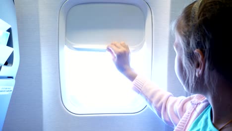 close-up.-kid-girl-lowers-the-porthole-curtain-in-the-airplane's-cabin,-from-there-shines-a-bright-light.-girl-looking-out-through-airplane's-window-viewing-Sky-and-Clouds-and-landscape-below