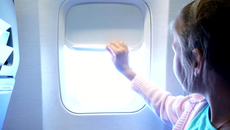 Close-up.-baja-de-niña-Kid-la-portilla-de-la-cortina-en-cabina-del-avión,-allí-brilla-una-luz-brillante.-chica-mirando-a-través-de-la-ventana-del-avión-viendo-el-cielo-y-las-nubes-y-el-paisaje-abajo