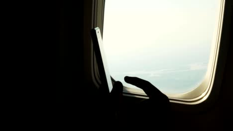 close-up.-dark-silhouette-of-kid-hands-and-mobile-phone-against-airplane's-illuminator.-Child-using,-plays-game-on-smartphone,-tablet-phone-in-airplane-cabin.-passengers-use-gadgets-during-the-flight.-Flight-entertainment-for-kids