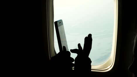 close-up.-dark-silhouette-of-kid-hands-and-mobile-phone-against-airplane's-illuminator.-Child-using,-plays-game-on-smartphone,-tablet-phone-in-airplane-cabin.-passengers-use-gadgets-during-the-flight.-Flight-entertainment-for-kids