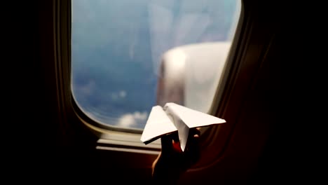 close-up.-Silhouette-of-a-child's-hand-with-small-paper-plane-against-the-background-of-airplane-window.-Child-sitting-by-aircraft-window-and-playing-with-little-paper-plane.-during-flight-on-airplane