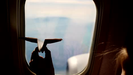 close-up.-Silhouette-of-a-child's-hand-with-small-paper-plane-against-the-background-of-airplane-window.-Child-sitting-by-aircraft-window-and-playing-with-little-paper-plane.-during-flight-on-airplane