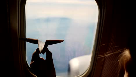 Close-up.-Silhouette-einer-Kinderhand-mit-kleinen-Papierflieger-vor-dem-Hintergrund-der-Flugzeugfenster.-Kind-Flugzeug-Fenster-sitzen-und-spielen-mit-kleinen-Papierflieger.-während-des-Fluges-im-Flugzeug