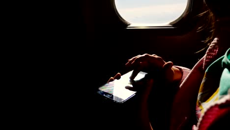 Close-up.-die-Sonnenstrahlen-spiegeln-sich-in-das-Telefon.-dunkle-Silhouette-Kind-Hände-und-Handy-gegen-Flugzeug-Hilfslicht.-Kind-mit,-spielt-Spiel-auf-Smartphone,-Tablet-Telefon-in-der-Flugzeugkabine