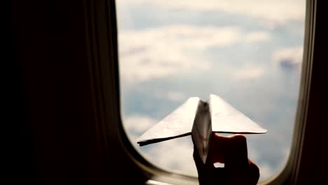 close-up.-Silhouette-of-a-child's-hand-with-small-paper-plane-against-the-background-of-airplane-window.-Child-sitting-by-aircraft-window-and-playing-with-little-paper-plane.-during-flight-on-airplane