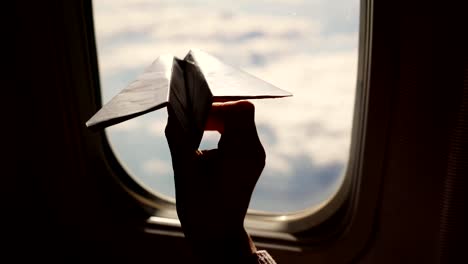 close-up.-Silhouette-of-a-child's-hand-with-small-paper-plane-against-the-background-of-airplane-window.-Child-sitting-by-aircraft-window-and-playing-with-little-paper-plane.-during-flight-on-airplane