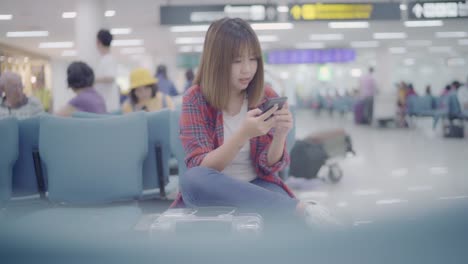 Happy-Asian-woman-using-and-checking-her-smartphone-while-sitting-on-chair-in-terminal-hall-while-waiting-her-flight-at-the-departure-gate-in-international-airport.-Women-happy-in-airport-concept.