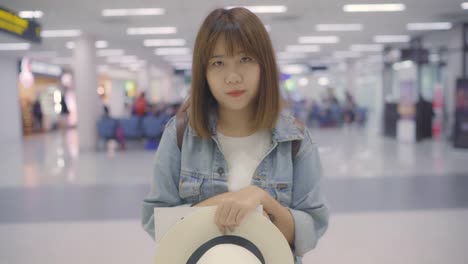 Happy-Asian-woman-smiling-to-camera-while-staying-at-terminal-in-international-airport,-female-tourist-ready-to-travel-on-vacation.-Lifestyle-women-happy-holiday-in-the-airport-concept.