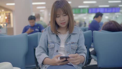 Happy-Asian-woman-using-and-checking-her-smartphone-while-sitting-on-chair-in-terminal-hall-while-waiting-her-flight-at-the-departure-gate-in-international-airport.-Women-happy-in-airport-concept.