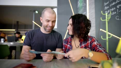 Happy-loving-couple-sitting-at-the-table-in-coffee-shop-and-looking-at-their-tickets.-Smiling-young-man-and-woman-are-looking-at-each-other,-examinating-their-boarding-passes-and-laughing