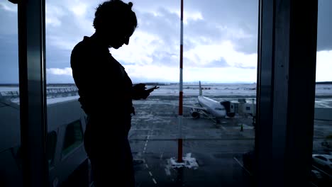 Silueta-de-forma-joven-de-pie-junto-a-la-ventana-de-aeropuerto-y-el-uso-de-su-smartphone.-Mujer-de-negocios-joven-bien-proporcionado-a-la-espera-de-su-embarque-cerca-de-la-puerta-con-la-ventana-y-el-plano-en-el-fondo