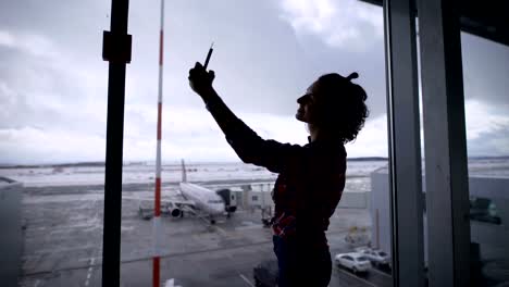 Hermosa-chica-morena-con-el-pelo-corto-en-el-aeropuerto.-Ella-es-feliz.-Puesto-que-se-iba-de-vacaciones-y-planes-de-viaje-es-de-buen-humor.-Ventana-panorámica-en-el-aeropuerto-de-pie-hace-selfie