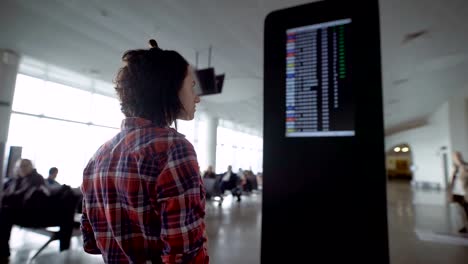 Niña-de-pie-en-el-aeropuerto-y-espera-anuncios-de-aterrizaje-a-bordo-de-la-aeronave.-Morena-teniendo-en-cuenta-el-marcador-con-un-programa-de-los-aviones,-mira-su-reloj-y-con-su-smartphone